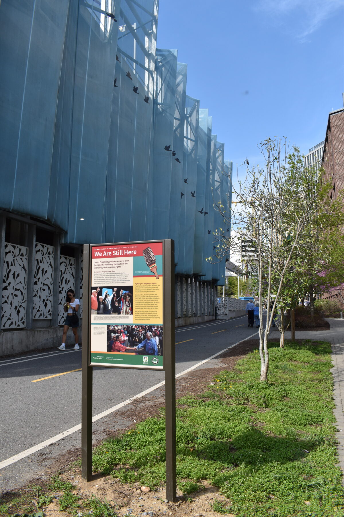 Color photo of a panel titled We Are Still Here next to a paved path and a tall building.