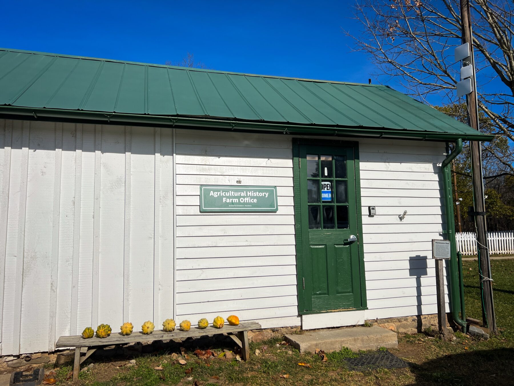 White shed with green roof and door with green sign that says "Agricultural History Farm Office" Theres an "Open, Come In" sign hanging on door.